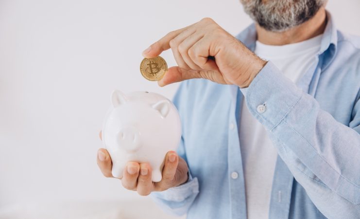 Eine Person hält ein weißes Sparschwein und legt eine physische Bitcoin-Münze hinein, was die Idee des Sparens mit einem Bitcoin-Sparplan symbolisiert.