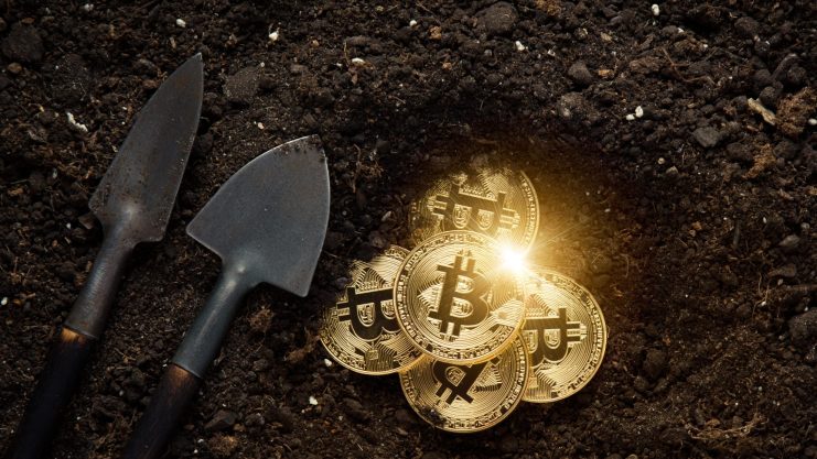 Zwei kleine Schaufeln liegen auf dem Boden neben einem Haufen goldfarbener Bitcoin-Münzen, die teilweise in der Erde vergraben sind und das Konzept des Mining von Kryptowährungen symbolisieren.