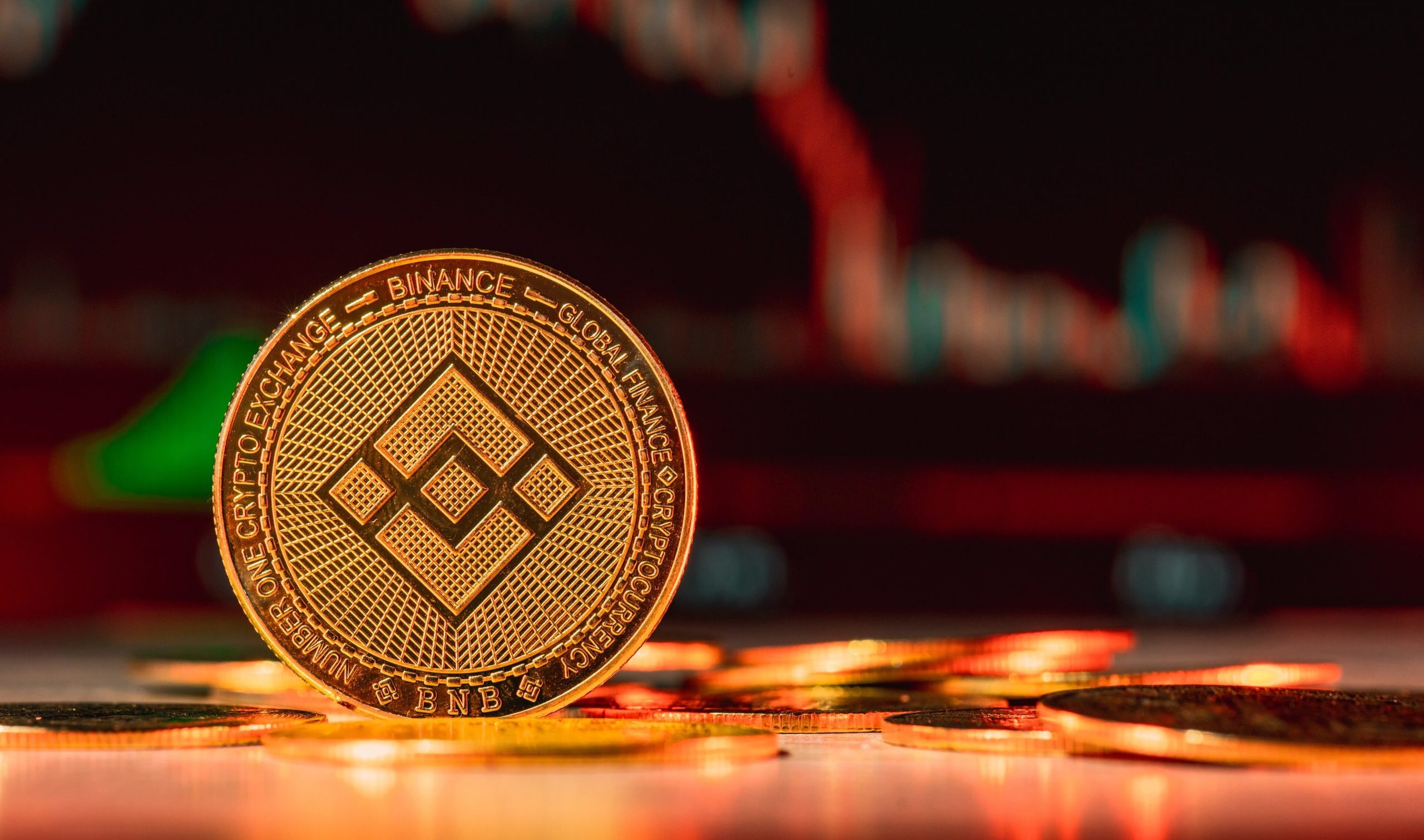 Eine goldene BNB Binance-Münze steht aufrecht auf einer Fläche mit mehreren anderen Münzen vor dem verschwommenen Hintergrund eines digitalen Finanzdiagramms.