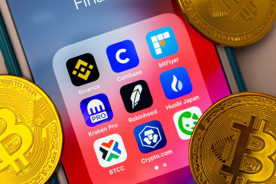 Smartphone-Bildschirm mit Symbolen verschiedener Kryptowährungshandels-Apps, einschließlich CEX Token, umgeben von physischen Bitcoin-Münzen.