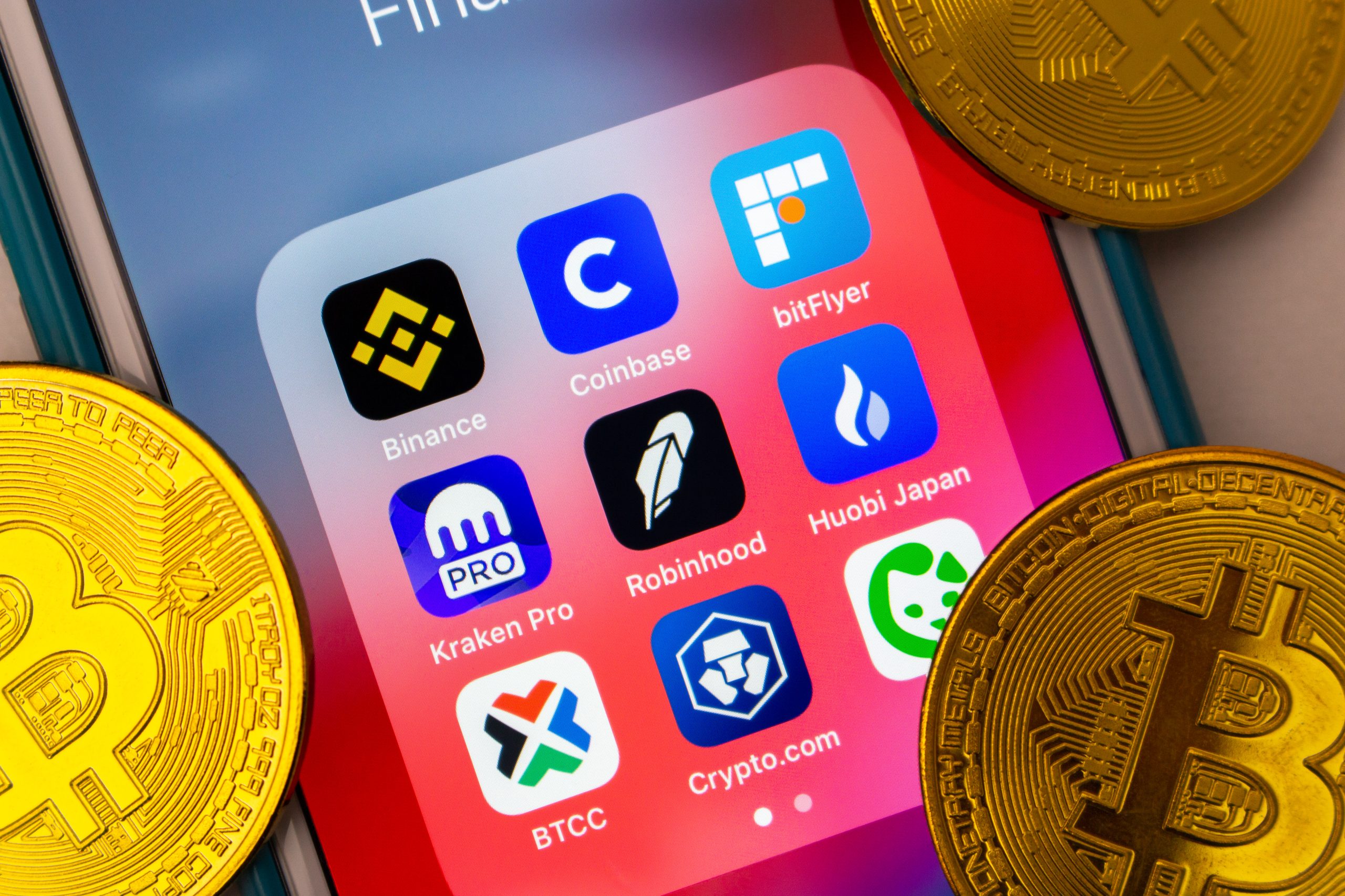 Smartphone-Bildschirm mit Symbolen verschiedener Kryptowährungshandels-Apps, einschließlich CEX Token, umgeben von physischen Bitcoin-Münzen.