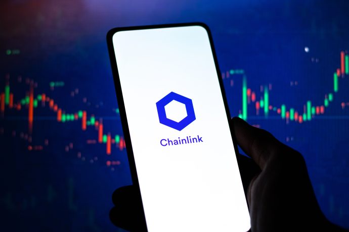 Eine Hand hält ein Smartphone, auf dem das Chainlink-Logo abgebildet ist, mit einem unscharfen Kryptowährungspreisdiagramm und Forex-Daten im Hintergrund.