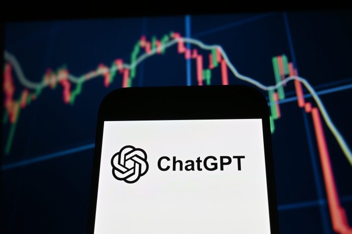 Ein Smartphone zeigt das ChatGPT-Logo und den Namen an, während im Hintergrund ein verschwommener Bitcoin-Candlestick-Chart zu sehen ist.