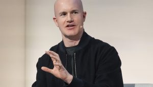 Ein glatzköpfiger Mann mit schwarzer Jacke und Coinbase-Headset-Mikrofon spricht, während er mit der Hand gestikuliert, vor einem schlichten Hintergrund.