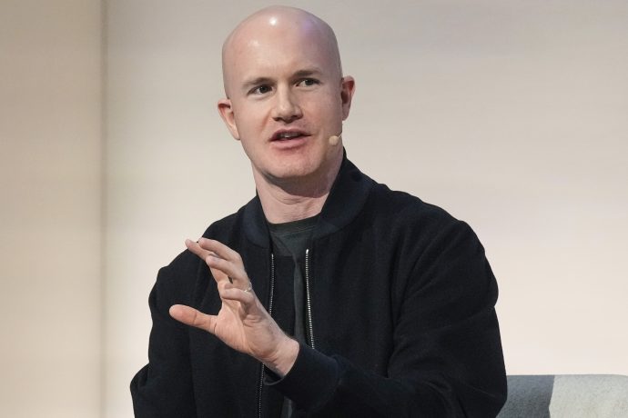 Ein glatzköpfiger Mann mit schwarzer Jacke und Coinbase-Headset-Mikrofon spricht, während er mit der Hand gestikuliert, vor einem schlichten Hintergrund.