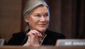 Eine Frau mit hellem Haar und einem dunklen Blazer (Cynthia Lummis) sitzt mit gefalteten Händen an einem Tisch und lächelt, während im Vordergrund ein verschwommenes Krypto-Schild zu sehen ist.