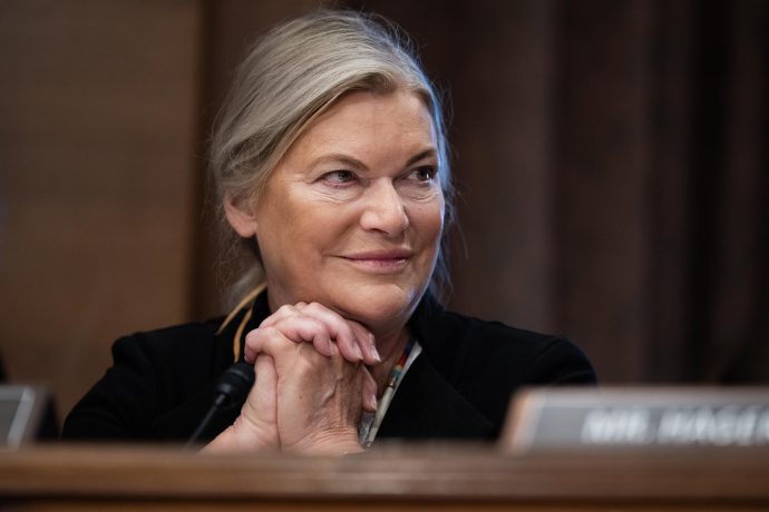 Eine Frau mit hellem Haar und einem dunklen Blazer (Cynthia Lummis) sitzt mit gefalteten Händen an einem Tisch und lächelt, während im Vordergrund ein verschwommenes Krypto-Schild zu sehen ist.