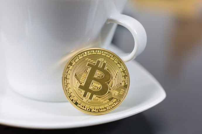 Eine goldfarbene Krypto-Bitcoin-Münze ruht aufrecht vor einer weißen Kaffeetasse mit Untertasse.