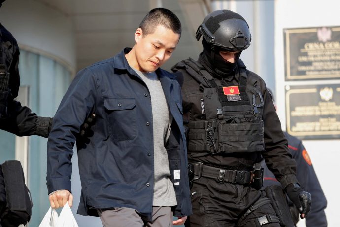 Do Kwon wird von einem bewaffneten Sicherheitsbeamten in taktischer Ausrüstung eskortiert; er hält eine weiße Plastiktüte in der Hand und schaut nach unten.