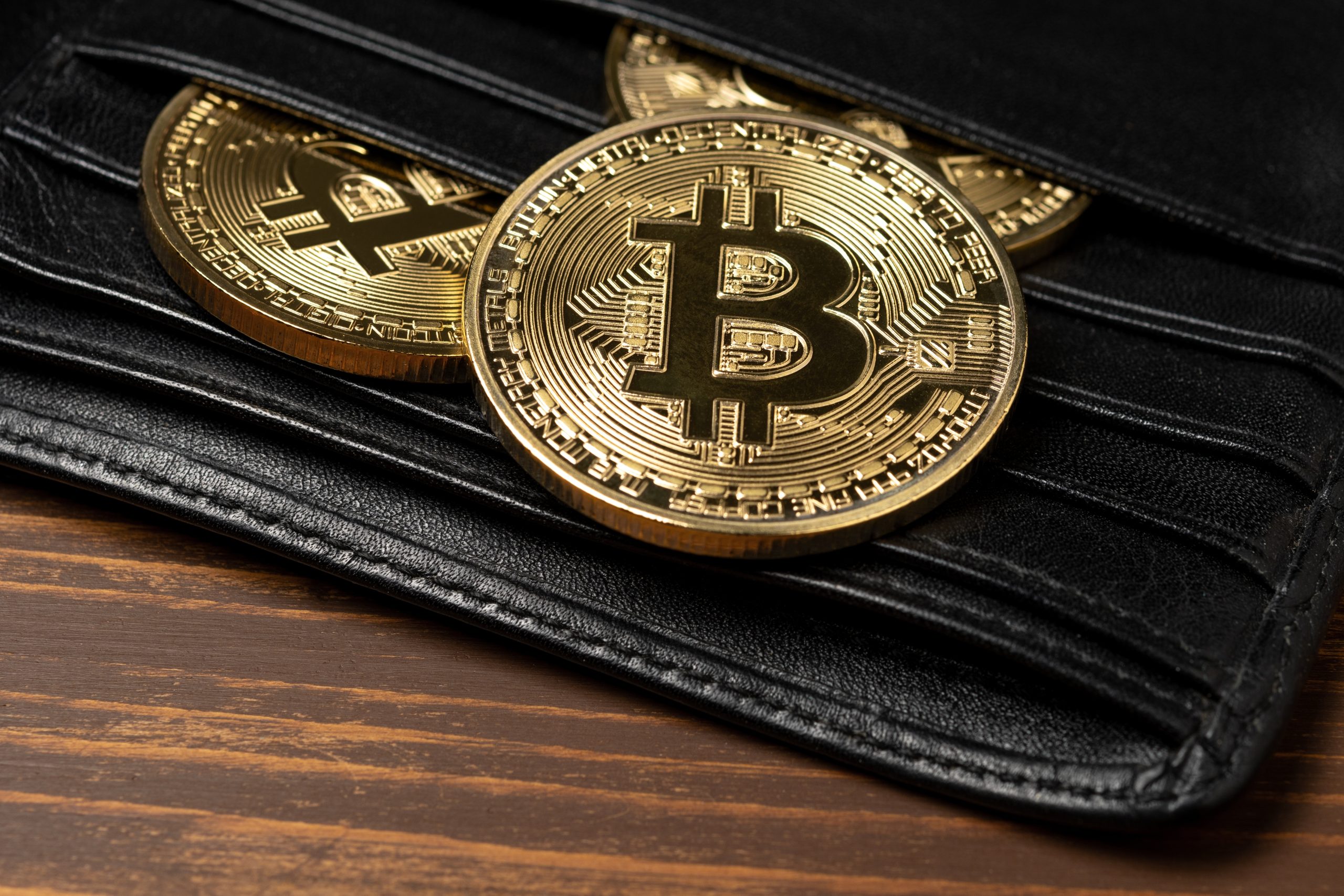 Drei goldfarbene Bitcoin-Münzen sind teilweise in einer schwarzen Ledergeldbörse auf einer hölzernen Oberfläche untergebracht, was die greifbare Darstellung von Bitcoin hervorhebt.