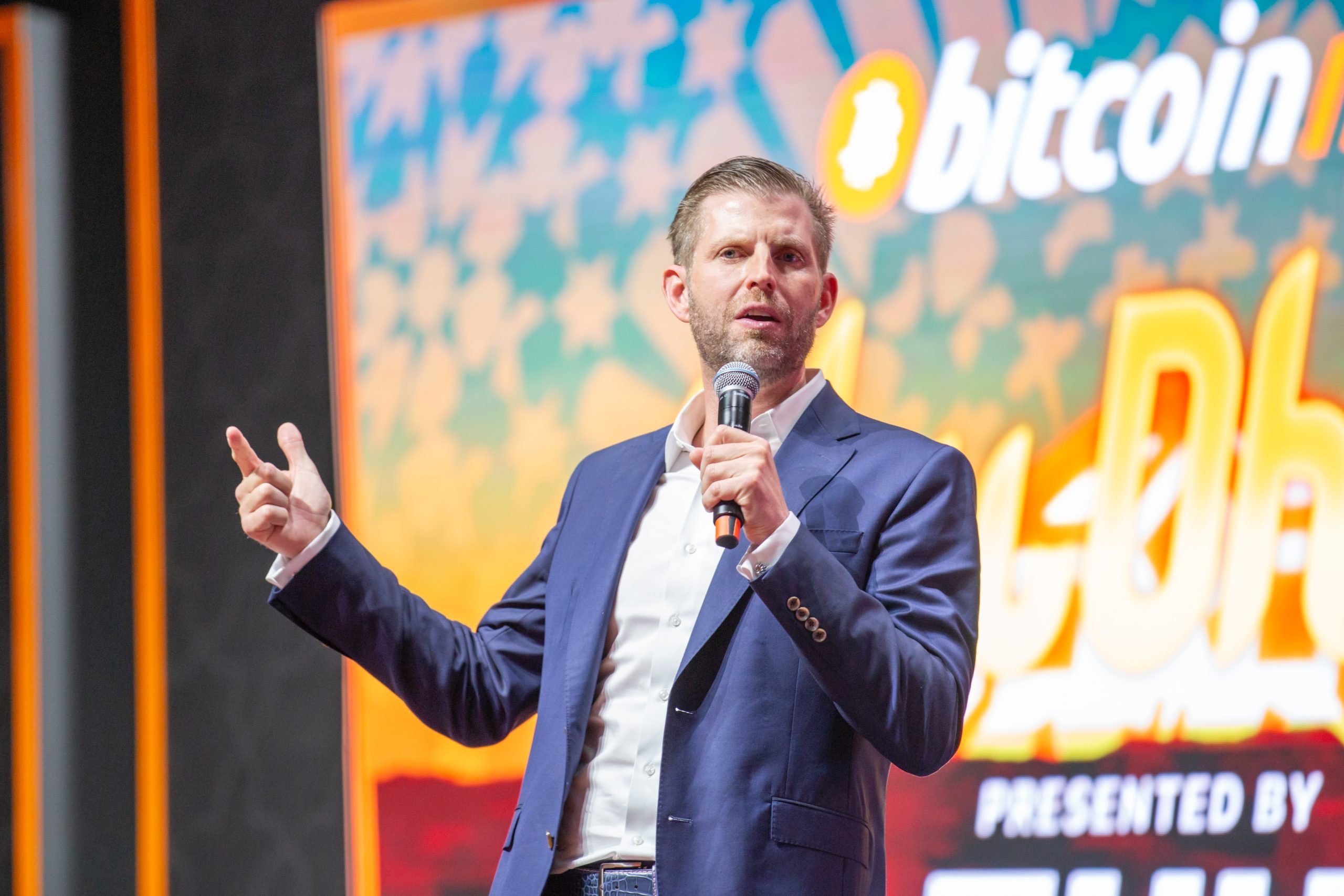 Ein Mann im Anzug spricht auf der Bühne vor einem großen Bitcoin-Konferenzschild in ein Mikrofon.