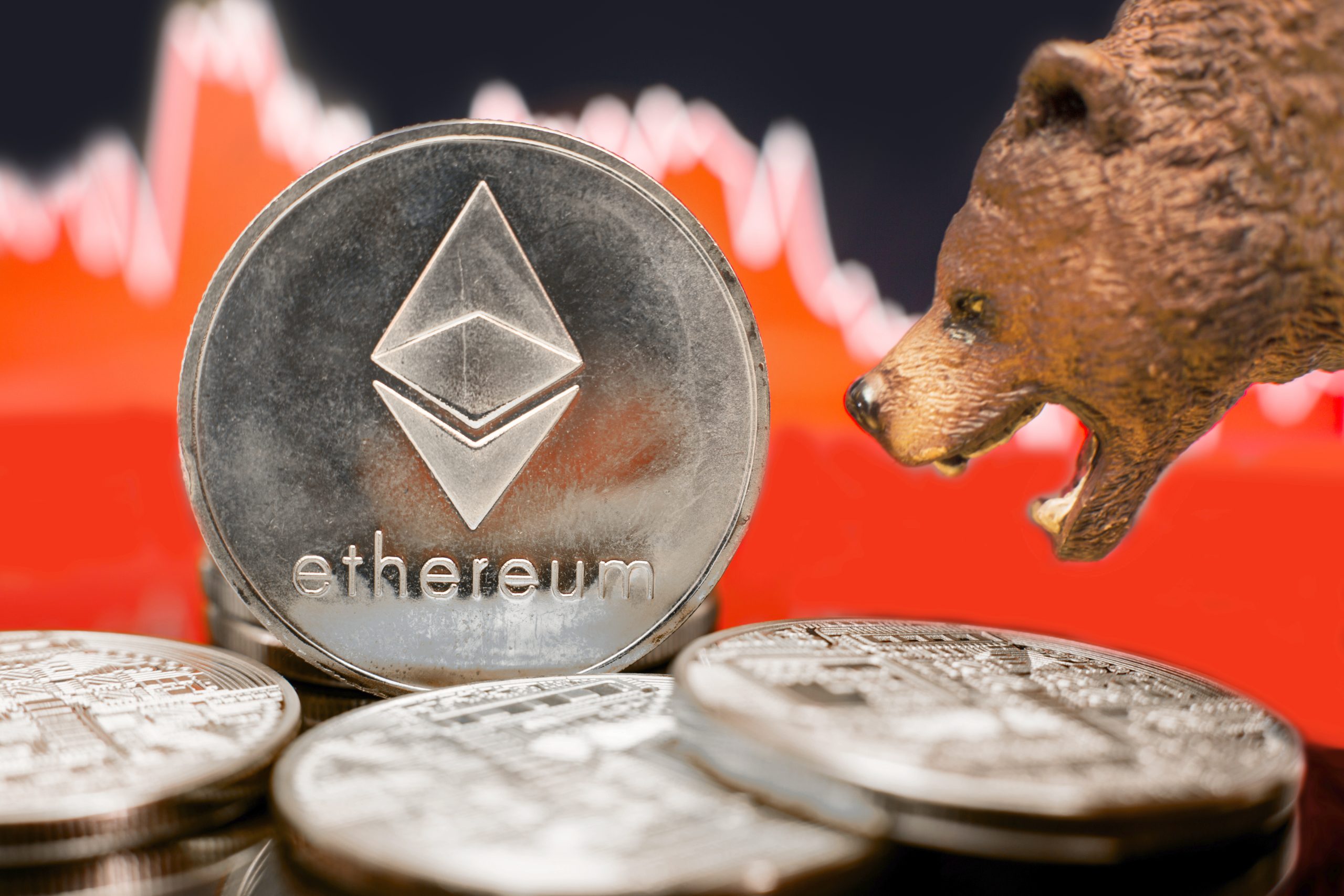 Eine Bärenfigur steht vor gestapelten Ethereum-Münzen mit einem roten Aktienchart im Hintergrund, der die Sorge vor einem ETH-Crash und einem potenziellen Abwärtstrend des Ethereum-Wertes hervorhebt.