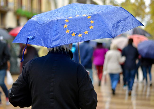 Eine Person hält einen blauen Regenschirm mit gelben EU-Sternen in der Hand, während sie an einem regnerischen Tag in einer Menschenmenge spazieren geht, als Symbol für die Hoffnung auf eine Krypto-Wende in Europa.