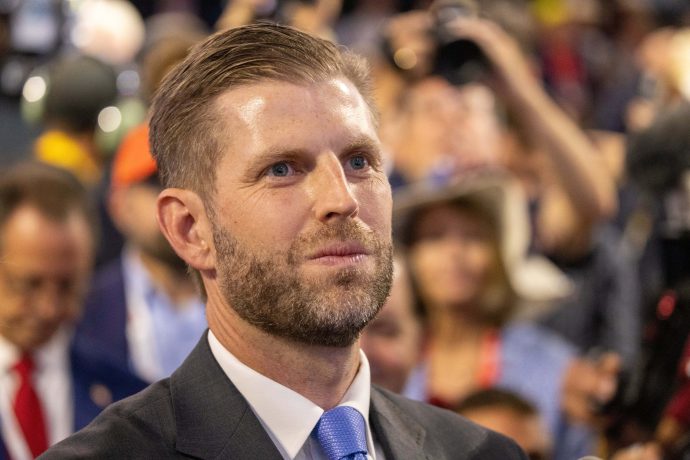 Ein Mann in Anzug und blauer Krawatte steht inmitten einer Menschenmenge und blickt nach vorne, was an die selbstbewusste Präsenz von Trump erinnert. Die Menschen im Hintergrund scheinen Fotos zu machen oder ein Ereignis zu beobachten.