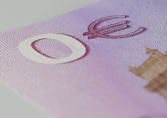 Nahaufnahme einer Ecke einer violetten Euro-Banknote, die einen Teil eines Gebäudes, das Euro-Währungssymbol und einen subtilen Hinweis auf Bitcoin zeigt.