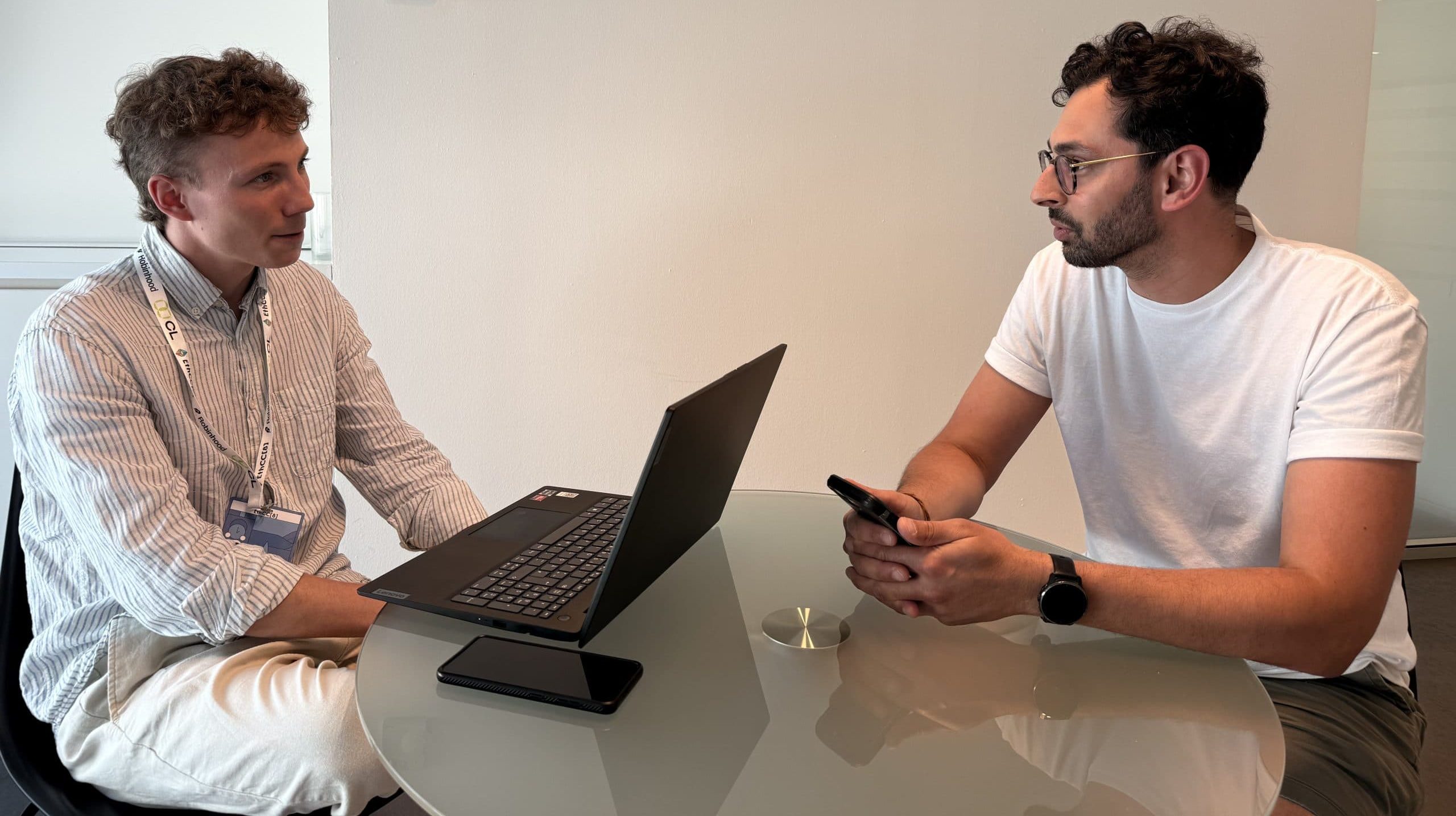 Zwei Männer (Johannes Dexl und Sebastien Gilquin) sitzen an einem runden Glastisch in einem modernen Büro, einer mit einem Laptop, der andere mit einem Smartphone in der Hand, und diskutieren über die neueste Hardware-Wallet-Technologie.