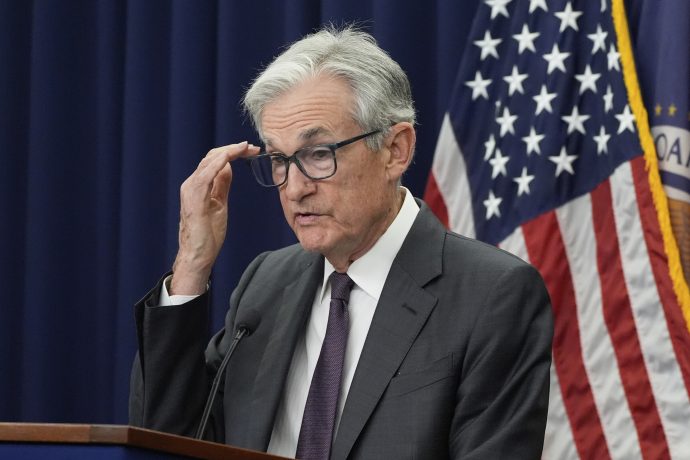 Ein Mann in Anzug und Krawatte (Jerome Powell) rückt seine Brille zurecht, während er auf einem Podium über Bitcoin spricht, mit der amerikanischen Flagge und einer weiteren Flagge im Hintergrund.
