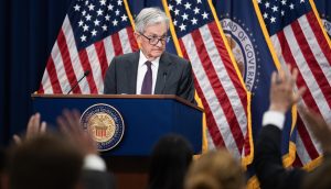 Jerome Powell steht an einem Podium mit dem Siegel der Federal Reserve vor mehreren amerikanischen Flaggen in Jackson Hole und wendet sich mit erhobenen Händen an das Publikum.