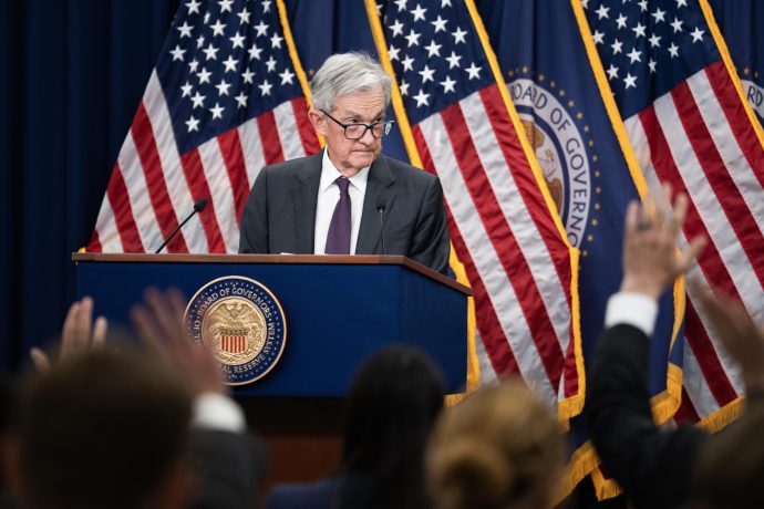 Jerome Powell steht an einem Podium mit dem Siegel der Federal Reserve vor mehreren amerikanischen Flaggen in Jackson Hole und wendet sich mit erhobenen Händen an das Publikum.