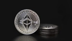 Eine silberne Ethereum-Münze steht aufrecht vor einem kleinen Stapel ähnlicher Ethereum-Münzen, die sich alle vor einem schwarzen Hintergrund befinden.