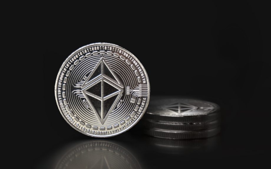 Eine silberne Ethereum-Münze steht aufrecht vor einem kleinen Stapel ähnlicher Ethereum-Münzen, die sich alle vor einem schwarzen Hintergrund befinden.