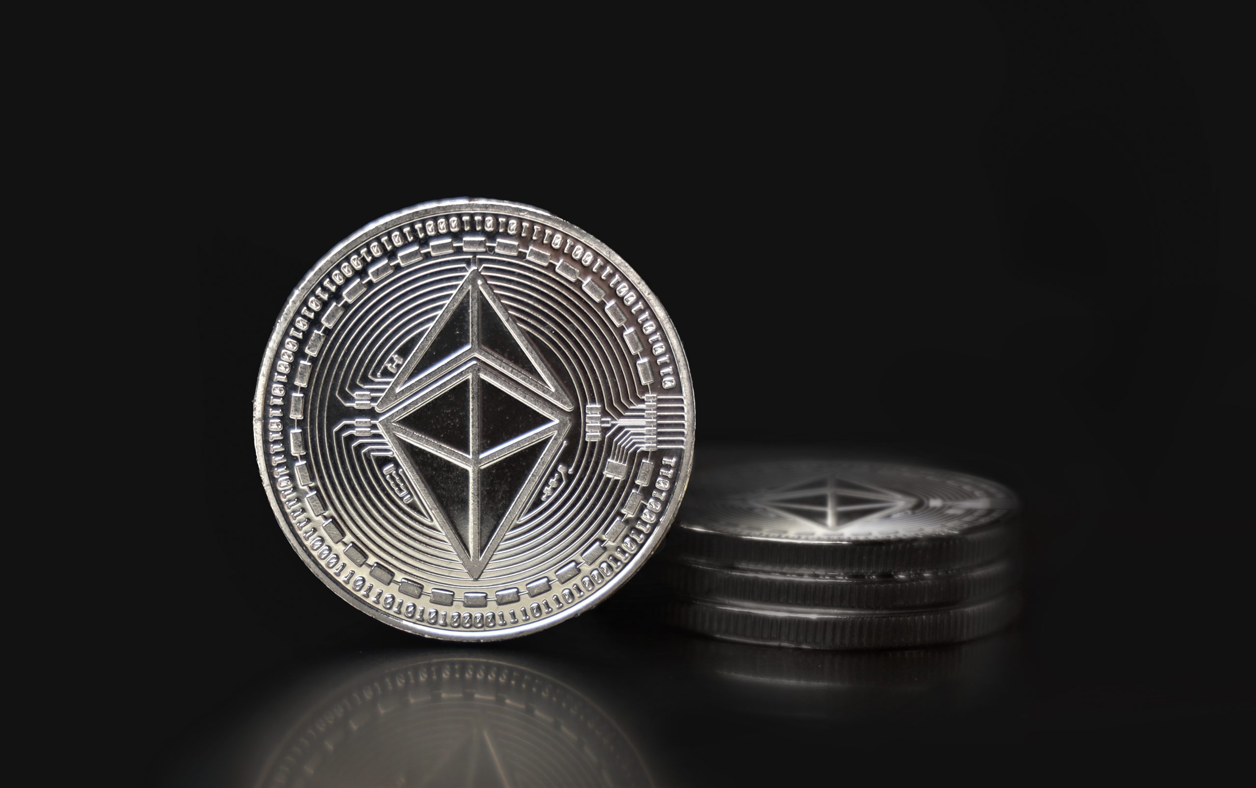 Eine silberne Ethereum-Münze steht aufrecht vor einem kleinen Stapel ähnlicher Ethereum-Münzen, die sich alle vor einem schwarzen Hintergrund befinden.