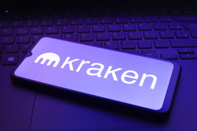 Ein Smartphone mit dem Kraken-Logo und dem Namen auf dem Bildschirm liegt auf einer Laptop-Tastatur, die von einem violetten Kraken-Glühen erleuchtet wird.