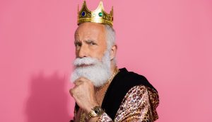 Älterer Mann mit weißem Bart, goldener Krone und luxuriösem Outfit vor pinkem Hintergrund – symbolisiert finanziellen Krypto-Erfolg und Selbstbewusstsein.