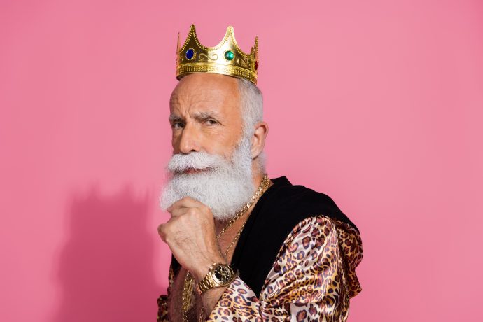 Älterer Mann mit weißem Bart, goldener Krone und luxuriösem Outfit vor pinkem Hintergrund – symbolisiert finanziellen Krypto-Erfolg und Selbstbewusstsein.