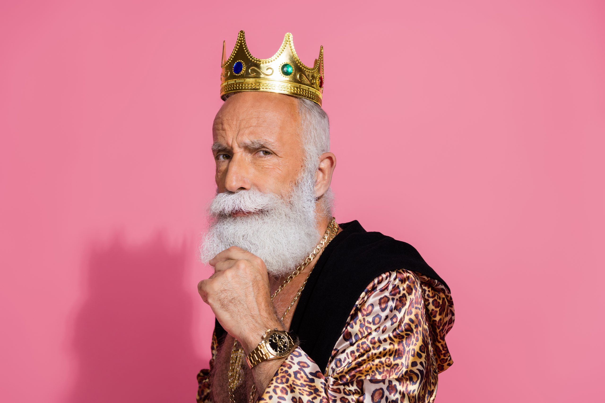 Älterer Mann mit weißem Bart, goldener Krone und luxuriösem Outfit vor pinkem Hintergrund – symbolisiert finanziellen Krypto-Erfolg und Selbstbewusstsein.