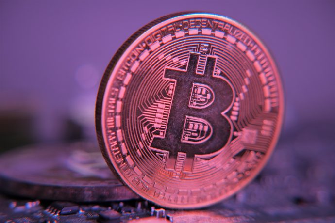 Eine Nahaufnahme eines physischen Bitcoin-Tokens, der aufrecht steht, mit von Krypto inspirierten Schaltkreisen und digitalen Mustern im Hintergrund bei violetter Beleuchtung.