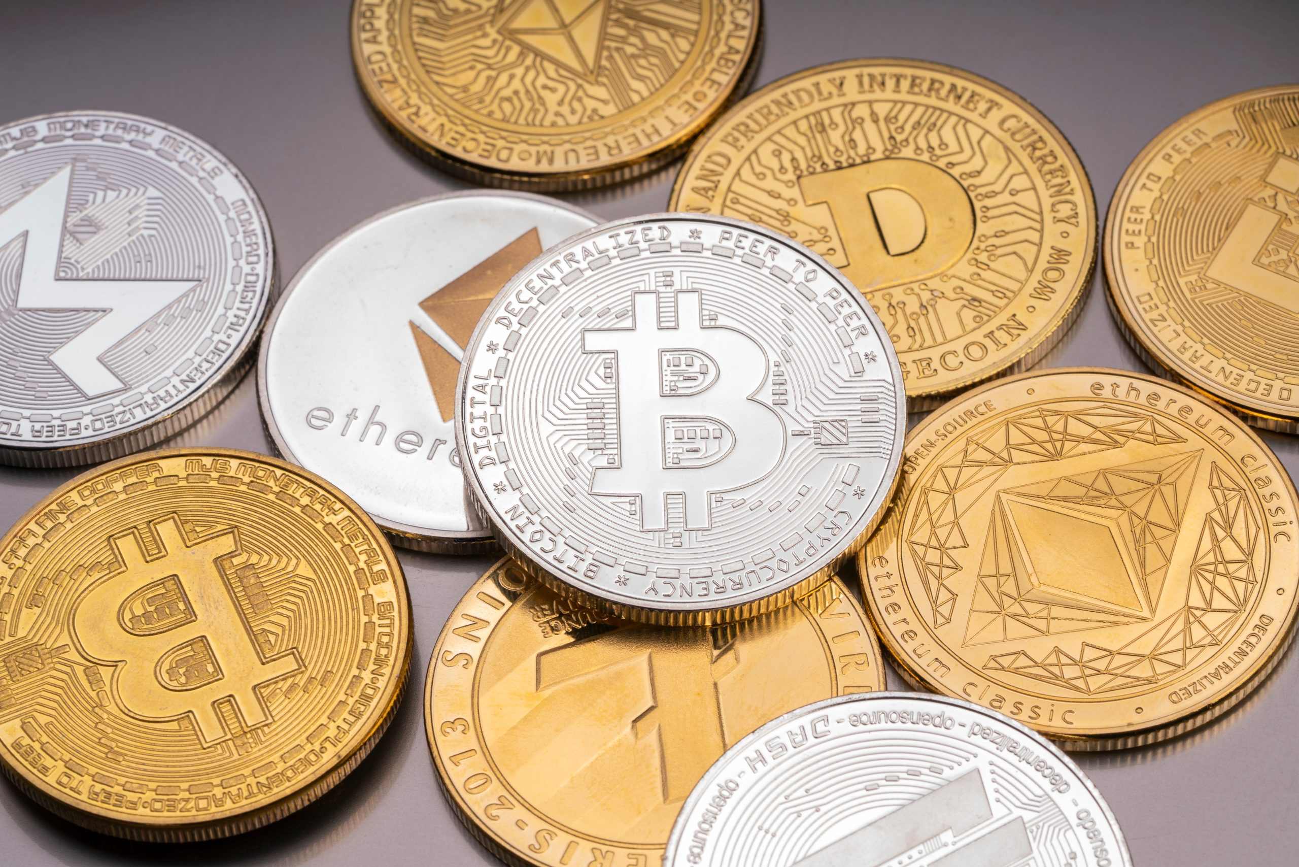 Verschiedene physische Krypto-Münzen, die verschiedene Kryptowährungen repräsentieren, darunter Bitcoin, Ethereum, Litecoin, Monero und Dogecoin, sind auf einer flachen Oberfläche verstreut.