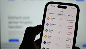 Eine Person hält ein Smartphone, auf dem Krypto-Kurse und Charts für Bitcoin, Ethereum, Tether, BNB, Solana und XRP angezeigt werden - alles in deutscher Sprache.