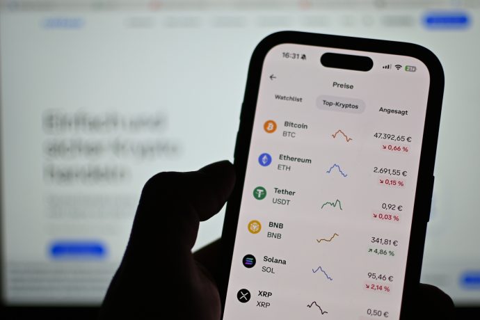 Eine Person hält ein Smartphone, auf dem Krypto-Kurse und Charts für Bitcoin, Ethereum, Tether, BNB, Solana und XRP angezeigt werden - alles in deutscher Sprache.