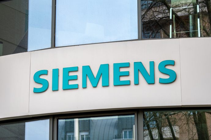 An der Außenfassade eines Gebäudes prangt das Siemens-Firmenlogo in fetten, türkisfarbenen Buchstaben über großen Fenstern, daneben eine schlichte Krypto-Beschilderung.