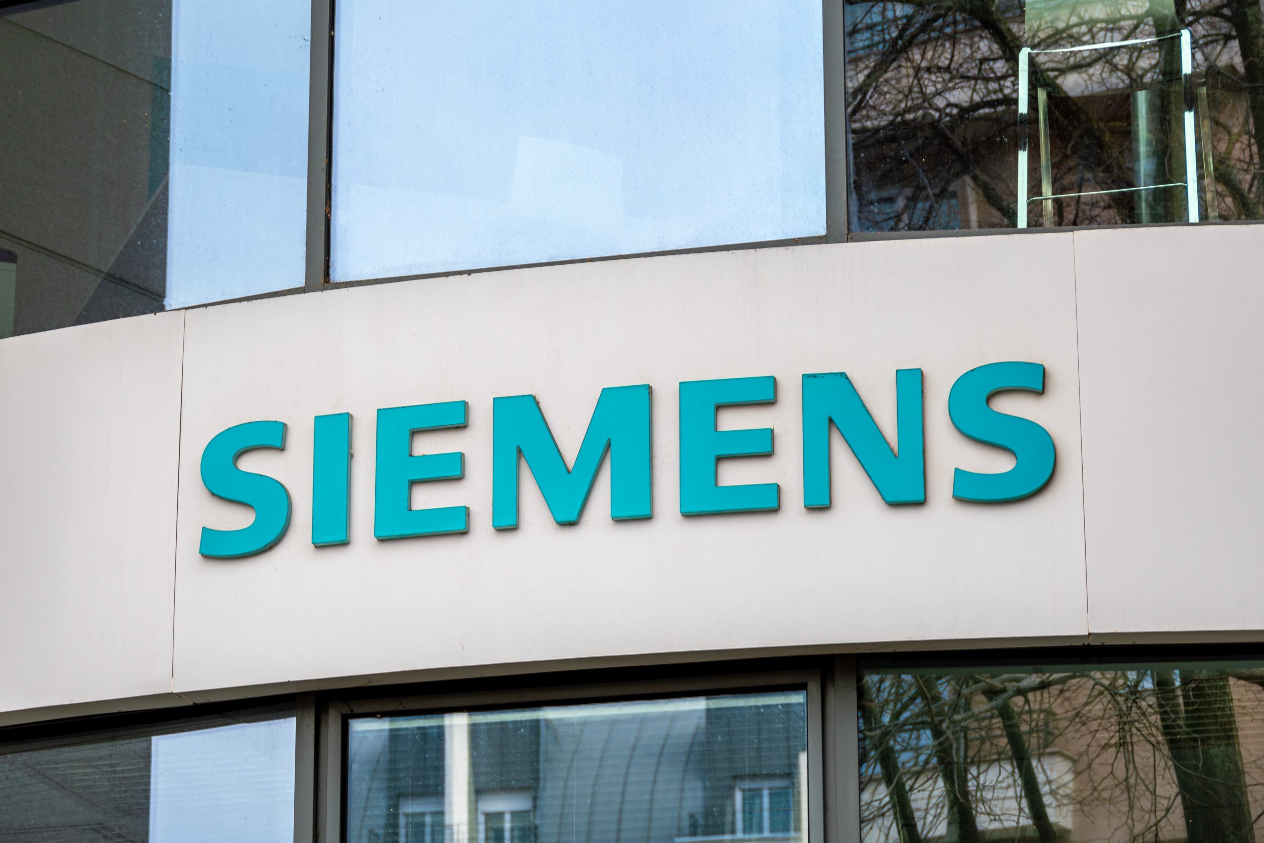 An der Außenfassade eines Gebäudes prangt das Siemens-Firmenlogo in fetten, türkisfarbenen Buchstaben über großen Fenstern, daneben eine schlichte Krypto-Beschilderung.