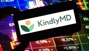 Eine Hand hält ein Gerät mit dem KindlyMD-Logo vor einem Bildschirm mit bunten Finanzcharts, Börsendaten und Bitcoin-Trends.