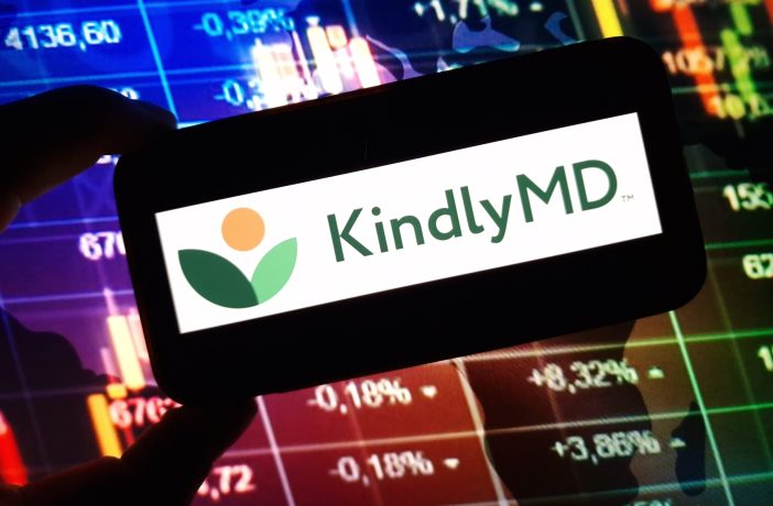 Eine Hand hält ein Gerät mit dem KindlyMD-Logo vor einem Bildschirm mit bunten Finanzcharts, Börsendaten und Bitcoin-Trends.