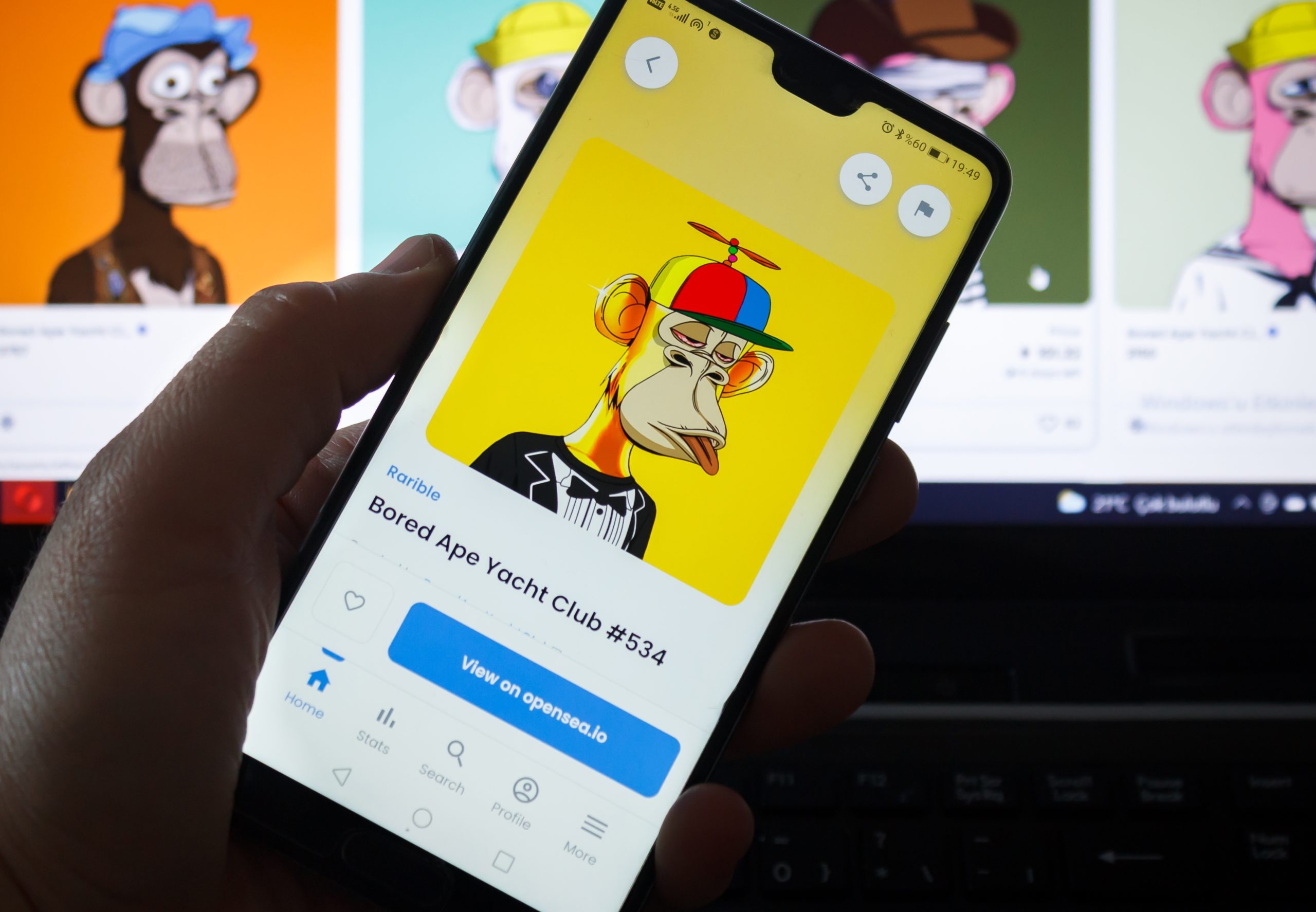 Eine Hand hält ein Smartphone, auf dem ein Bored Ape Yacht Club NFT mit gelbem Hintergrund und Propellerhut zu sehen ist, während auf einem Computerbildschirm ähnliche Bilder erscheinen, die die NFT-Handelstrends in Deutschland hervorheben.