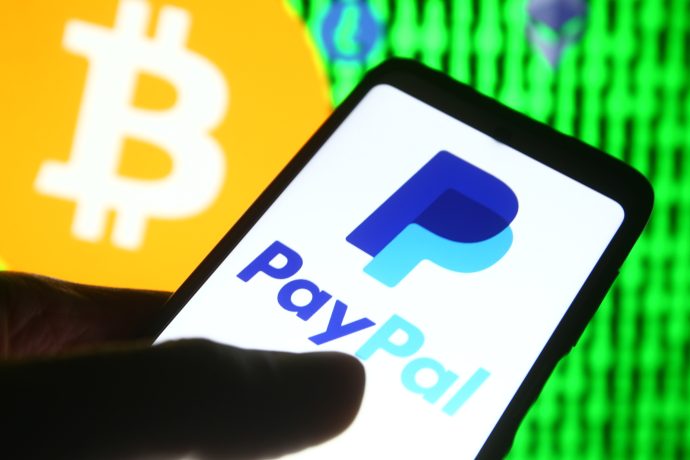 Ein Smartphone zeigt im Vordergrund das PayPal-Zahlungs-Logo, dahinter ist ein großes Bitcoin-Symbol und ein verpixelter grüner Hintergrund zu sehen.