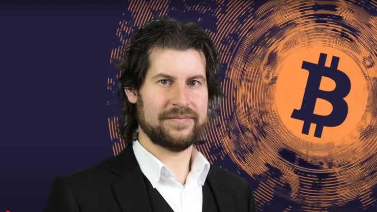 Ein Mann im Anzug steht selbstbewusst vor einem grafischen Hintergrund, der ein großes Bitcoin-Symbol zeigt. Es ist Rahim Taghizadegan.