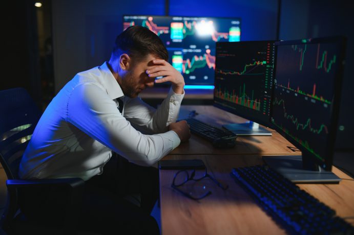 Ein Mann in Hemd und Krawatte sitzt an einem Schreibtisch mit mehreren Monitoren, auf denen Aktien- und Bitcoin-Charts zu sehen sind, und stützt seinen Kopf in die Hand, er wirkt verzweifelt.