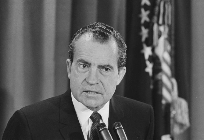 Ein Mann im Anzug spricht an einem Podium mit Mikrofonen, mit einer verschwommenen US-Flagge im Hintergrund, die an Richard Nixons Rede vor 54 Jahren erinnert, in der er das Ende des Goldstandards ankündigte.