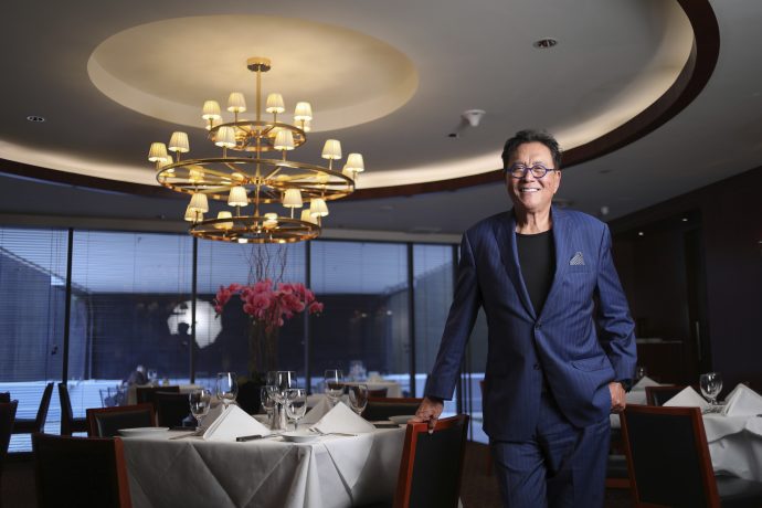 Ein Mann in einem blauen Anzug steht lächelnd in einem eleganten Restaurant mit gedeckten Tischen, großen Fenstern und einem Kronleuchter über dem Kopf und feiert einen kürzlich erzielten Bitcoin-Erfolg.