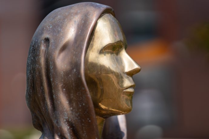 Seitenansicht einer Metallstatue, die Satoshi Nakomoto mit einem menschlichen Gesicht mit Kapuze darstellt, mit einer glatten, reflektierenden goldenen Oberfläche und einer bronzefarbenen Kapuze.