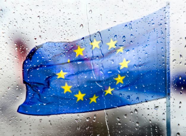 Die Flagge der Europäischen Union mit gelben Sternen auf blauem Grund, gesehen durch ein regenverhangenes Fenster mit Wassertropfen, erinnert an die Ungewissheit der Kryptomärkte inmitten sich ändernder Vorschriften.