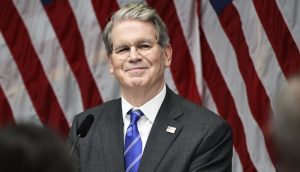 Ein Mann in Anzug und Krawatte (US-Finanzminister Scott Bessent) steht vor mehreren amerikanischen Flaggen und lächelt von einem Podium mit einem Mikrofon, als der US-Finanzminister neue Bitcoin-Regelungen ankündigt.