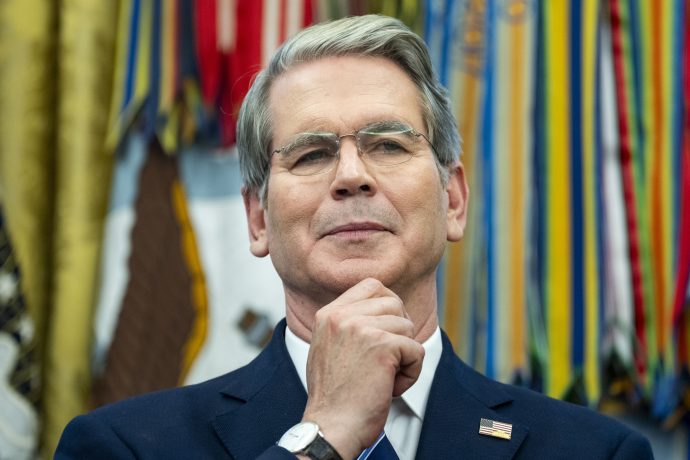 Ein Mann im Anzug mit einer Anstecknadel mit der amerikanischen Flagge, einer Brille und einer Bitcoin-Anstecknadel steht vor bunten Militärbändern und hält sich nachdenklich das Kinn.