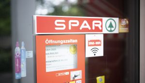 Am Eingang eines SPAR-Marktes in der Schweiz sind die Öffnungszeiten, das SPAR-Logo, ein WiFi-Schild und verschiedene Informationsaufkleber an der Glastür angebracht.