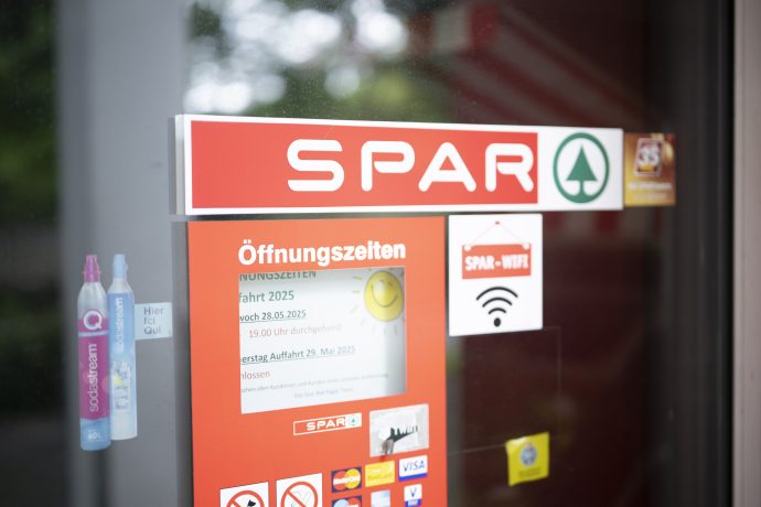 Am Eingang eines SPAR-Marktes in der Schweiz sind die Öffnungszeiten, das SPAR-Logo, ein WiFi-Schild und verschiedene Informationsaufkleber an der Glastür angebracht.